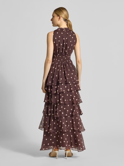 Modelka kobieca ma na sobie 'Abendkleid im Stufen-Look' od Neo Noir, brązowa z białymi kropkami, stufenowy fason maxi, krój taliowany, okrągły dekolt, kluczowy dekolt na plecach, długość tyłu w rozmiarze 36: 141 cm, szerokość w rozmiarze 36: 42 cm, widok całej sylwetki z tyłu.