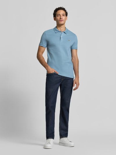 Männermodel trägt slim fit poloshirt aus puur katoen van Lacoste in lichtblauw, gecombineerd met donkere jeans, ganzkörperansicht van voren. Heren poloshirt van Lacoste