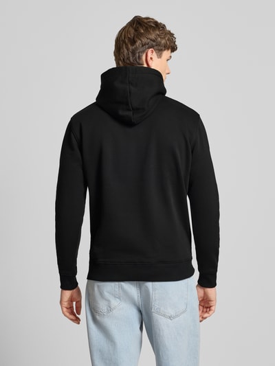 Männermodel trägt Hoodie mit Label-Print von Alpha Industries in Schwarz aus Baumwollmischung, lockere Passform, Unifarbenes-Design, elastische Abschlüsse, Kapuze mit Tunnelzug, Känguru-Tasche, Label-Details, Rückenlänge 70 cm, Ärmellänge 63 cm, Ganzkörperansicht Rückansicht.