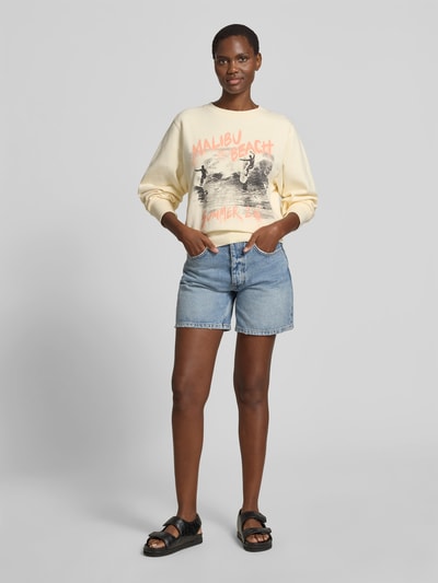 Frauenmodel trägt Relaxed Fit Sweatshirt mit Foto-Print von Anine Bing aus reiner Baumwolle mit Statement-Print, elastischen Abschlüssen, Rückenlänge bei Größe S 56 cm, hellgelbe Farbe, Motiv vorne, Ganzkörperansicht von vorne.