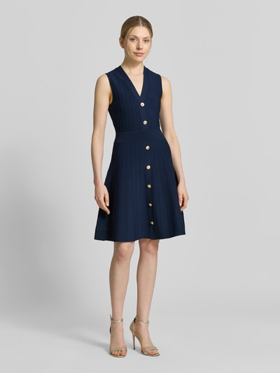 Frauenmodel trägt Knielanges Kleid aus Viskose-Mix Modell 'Amber' von Guess in Dunkelblau mit goldfarbenen Knöpfen, Strick-Optik, taillierte Passform, ärmellos, V-Ausschnitt, knielang; Ganzkörperansicht von vorne.