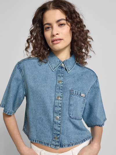 Frauenmodel trägt Cropped Hemdbluse in Denim-Optik von Calvin Klein Jeans, kurzärmelig, reine Baumwolle, Knopfleiste, Brusttasche, Nahaufnahme Oberkörper.