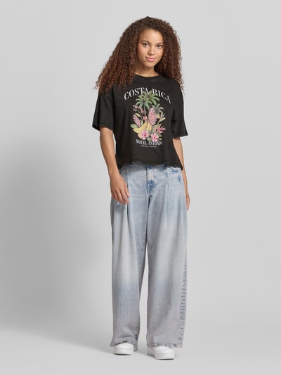 Frauenmodel trägt Damen T-Shirt von Only, Modell 'Boxy Fit T-Shirt aus reiner Baumwolle Modell LUCY' mit Optik: Unifarben, Boxy-Fit, Motiv-Print auf der Vorderseite, Kurzarm, Rundhals, Material: Reine Baumwolle, Hauptfarbe Schwarz mit buntem Print, glatte, weiche Textur, Ganzkörperansicht von vorne.