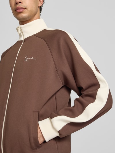 Männermodel trägt Sweatjacke mit Stehkragen von KARL KANI in Braun mit Creme, Material-Mix, Reißverschluss, Label-Stitching, Raglanärmel, Seitentaschen, elastische Abschlüsse, lockere Passform, Nahaufnahme Oberkörper.