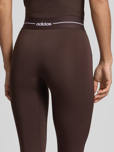 Frauenmodel trägt Regular Fit Leggings mit Label Print von adidas Sportswear in Braun mit High Waist und elastischem Bund, Detailaufnahme Rückansicht.