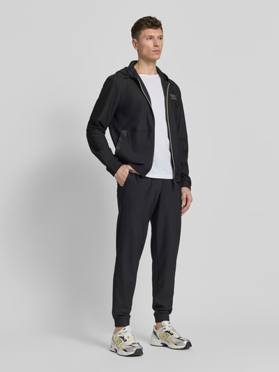 Mężczyzna nosi Regular Fit Sweatpants mit Strukturmuster von EA7 Emporio Armani, ciemny kolor, elastyczny materiał, kieszenie, widok całej sylwetki z przodu.
