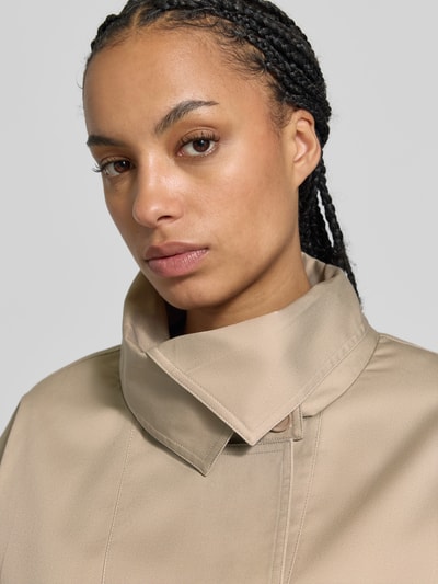 Vrouwenmodel draagt Kurzer Trenchcoat met katoenmix van ROTATE in beige, materiaalmix, getailleerde pasvorm, detailopname bovenlichaam. Context: dames trenchcoat, dubbele knoopsluiting, tailleband, verstelbare mouwsluitingen, ruglengte maat 36: 69 cm, borstwijdte 53 cm, mouwlengte 58 cm.