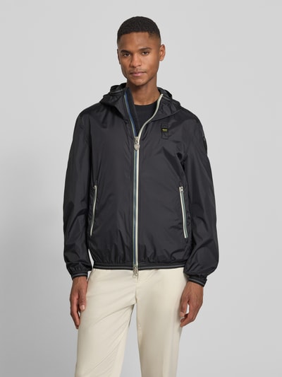 Männermodel trägt schwarze Jacke mit Label-Patches Modell 'Dodge' von Blauer Usa, Gerader-Schnitt, Kapuze, Ganzkörperansicht von vorne.