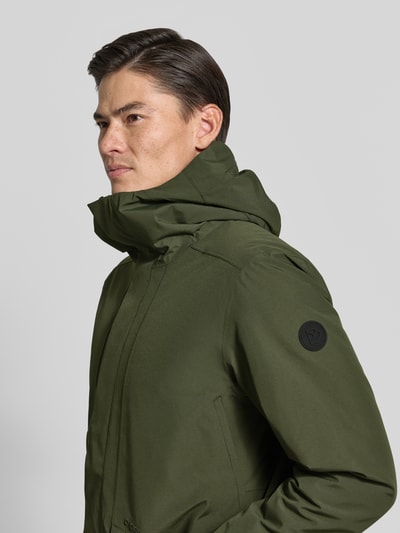 Männermodel trägt Regular Fit Jacke mit Kapuze Modell 'Adam' von Didriksons in Olivgrün, glatte Textur, Nahaufnahme Oberkörper. Rückenlänge, Schulterbreite, Brustweite und Ärmellänge bei Größe M laut Kontext.