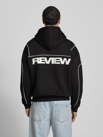 Męski model nosi Hoodie mit Logo und Känguru-Tasche od REVIEW, czarny z białym logo, krój regular fit, bawełniany mix, długość tyłu 71 cm, długość rękawa 67 cm, widok całej sylwetki z tyłu.