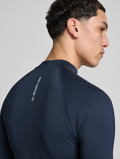Mannenmodel draagt donkerblauw Sonnenschutz Wet Shirt Surfshirt met UPF50 model 'EVERYDAY' van Quiksilver, labelprint, halflange mouwen, elastische materiaalmix, opstaande kraag, ruglengte bij maat M 71 cm, detailopname rug.