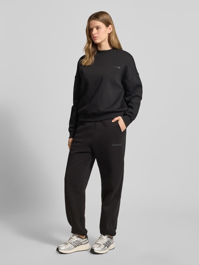 Frauenmodel trägt Basic Sweatpant mit Bündchen am Saum und seitlichen Taschen von Review, schwarze Farbe, Baumwoll-Mix, lockere Passform, Logo-Stickerei, Label-Print, Seitentaschen, elastische Abschlüsse, Ganzkörperansicht von vorne.