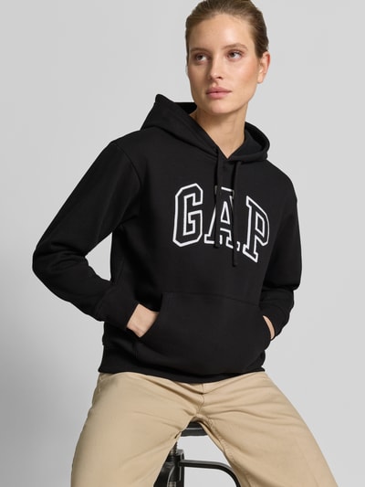 Kobiece model prezentuje Hoodie mit Logo und Känguru-Tasche od GAP, czarny, bawełniany krój regular fit, z logo i kieszenią kangurką, zdjęcie całej sylwetki z przodu.