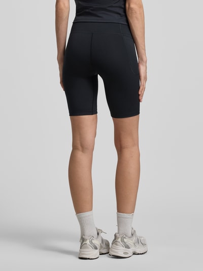 Frauenmodel trägt Radlerhose mit Logo von Under Armour in Schwarz, Material-Mix mit Stretch-Anteil, körpernahe Passform, elastischer Bund, Ganzkörperansicht Rückansicht.