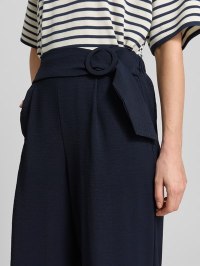 Vrouwenmodel draagt Wide Fit Stoffhose mit Bundfalten van Zero in donkerblauw met bandplooien, siergesp en verkorte pasvorm, detailopname taille.