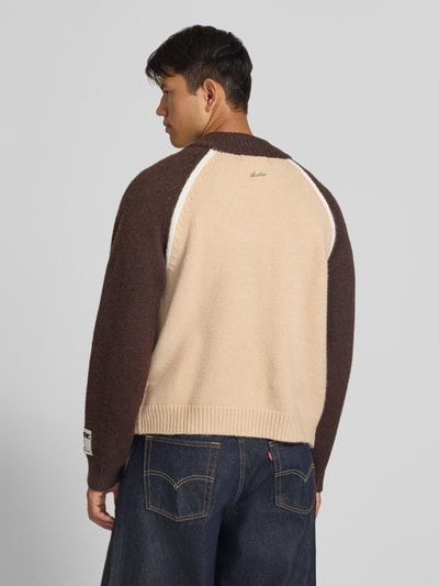 Männermodel trägt Strickjacke mit Label-Details von ANOTHER COTTON LAB, braune und beige Farbfelder, gerippte Abschlüsse, Rückenansicht, Ganzkörperansicht Rückansicht.