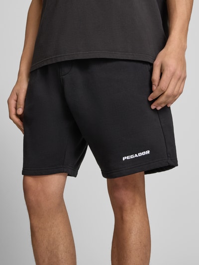 Männermodel trägt Shorts mit elastischem Bund und Logo-Stitching von Pegador aus Baumwoll-Mix, schwarze Farbe, Nahaufnahme Oberschenkel.