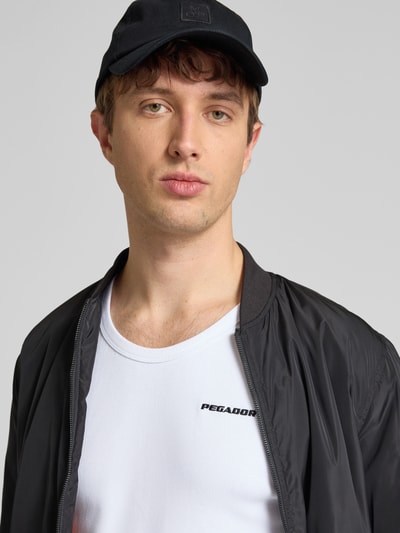 Męski model nosi top 'Tanktop mit Logo-Stitching und Rundhalsausschnitt' od Pegador, mieszanka bawełny z dodatkiem streczu, dopasowany krój, drobne prążkowanie, okrągły dekolt, wyhaftowane logo, długość tyłu w rozmiarze M: 69 cm, zdjęcie zbliżenie na górną część ciała.