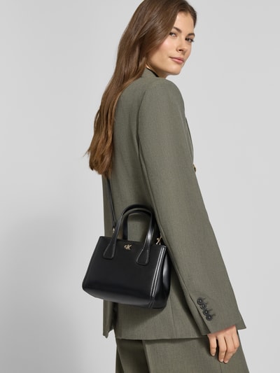 Kobiece model nosi czarną Torebka mit Label-Applikation von CK Calvin Klein, odpinany pasek na ramię, zapięcie na magnes, Ganzkörperansicht Rückansicht. Szerokość w rozmiarze 1: 18 cm, długość w rozmiarze 1: 14 cm, głębokość w rozmiarze 1: 9 cm, długość paska na ramię w rozmiarze 1: 108 cm.