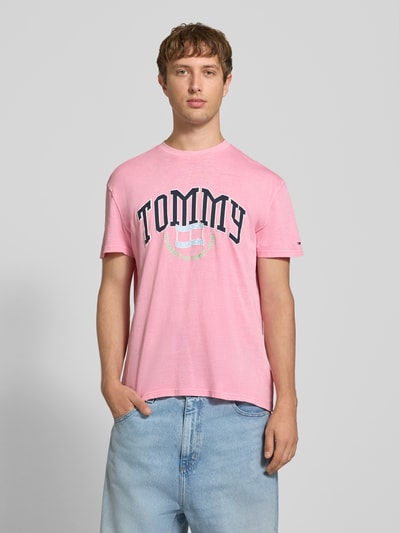 Männermodel trägt Regular Fit T-Shirt aus reiner Baumwolle von Tommy Jeans in Rosa mit rundem Schriftzug, Ganzkörperansicht von vorne.