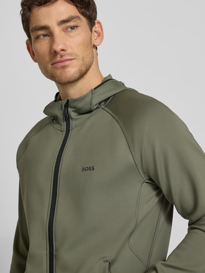 Männermodel trägt Regular Fit Trainingsjacke aus schnell trocknendem Gewebe Modell 'SICON ACTIVE' von BOSS Green, Optik: Unifarben, Kapuze, Langarm mit Daumenschlaufen, Reißverschluss, Label-Print, Reißverschlusstaschen, Nahaufnahme Oberkörper.