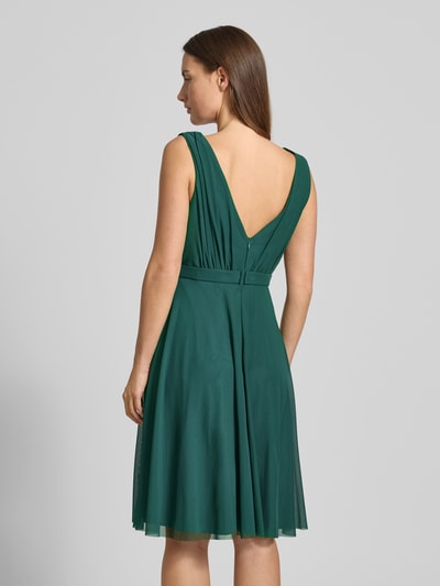Frauenmodel trägt Cocktailkleid mit V-Ausschnitt von TROYDEN COLLECTION in Dunkelgrün, taillierter Schnitt, Nahtreißverschluss, Ganzkörperansicht Rückansicht, Kontext: blickdichtes Unterkleid, Taillengürtel, Raffungen, Rückenlänge 79 cm, Brustweite 24 cm.