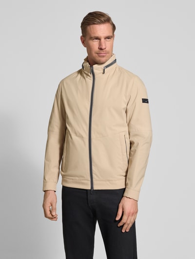 Männermodel trägt Regular Fit Jacke mit Stehkragen von bugatti in Beige mit Zweiwege-Reißverschluss, Eingrifftaschen, Logo-Detail, Ganzkörperansicht von vorne. Rückenlänge 68 cm, Schulterbreite 42 cm, Brustweite 50 cm, Ärmellänge 65 cm, verstaubare Kapuze, elastische Ärmelabschlüsse.
