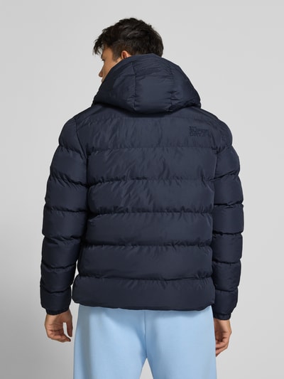 Männermodel trägt Steppjacke mit Label-Stitching und Kapuze von Superdry in Dunkelblau, Rückenansicht, Ganzkörperansicht Rückansicht. Gerader Schnitt, Kapuze mit Kordelzug, Reißverschluss, seitliche Eingrifftaschen, elastische Ärmelabschlüsse, strukturierte Steppung, Rückenlänge 67 cm, Schulterbreite 48 cm, Brustweite 58 cm, Ärmellänge 72 cm.