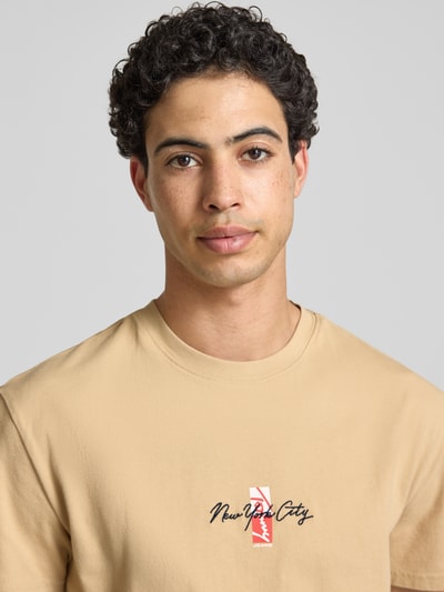 Männermodel trägt Regular Fit T-Shirt aus reiner Baumwolle von Tommy Jeans in Beige, Motiv-Print, Rundhals, Jersey, Nahaufnahme Oberkörper.
