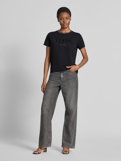 Damesmodel draagt t-shirt uit puur katoen met logodetail van GUESS, getailleerde pasvorm, met pailletten, ronde hals, ruglengte maat S 58 cm, zwart, rechte jeans, sandalen, ganzebodyaanzicht van voren.