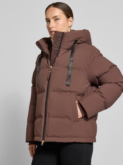 Kobieta nosi kurtkę w kolorze brązowym z efektem pikowania, wpuszczane kieszenie, kaptur z tunel, zamek błyskawiczny. Kurtka w stylu krój regular fit Model 'Jilly Hope' od MOS MOSH, Długość tyłu w rozmiarze S: 63 cm, szerokość ramion w rozmiarze S: 50 cm, szerokość klatki piersiowej w rozmiarze S: 58 cm, długość rękawów w rozmiarze S: 64 cm, wpuszczane kieszenie. Ganzkörperansicht z przodu.
