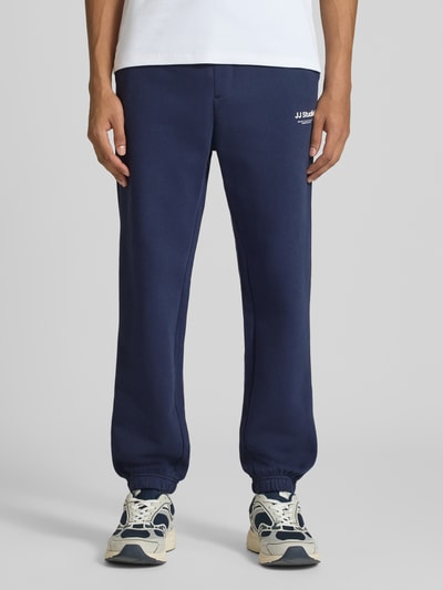 Männermodel trägt dunkelblaue Straight Fit Sweatpants aus Baumwoll-Mix Modell 'Kane' von JACK & JONES mit Logo-Detail, Nahaufnahme Unterkörper. Herren Sweatpants, elastischer Bund und Beinabschlüsse, Gesäßtasche, neutraler Hintergrund, Kontext: Innenbeinlänge bei Größe M 72 cm.