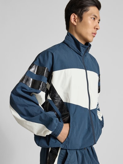 Männermodel trägt Trainingsjacke mit Stehkragen von VETEMENTS, Farbkombination Blau, Weiß, Schwarz, Zweiwege-Reißverschluss, Nahaufnahme Oberkörper.