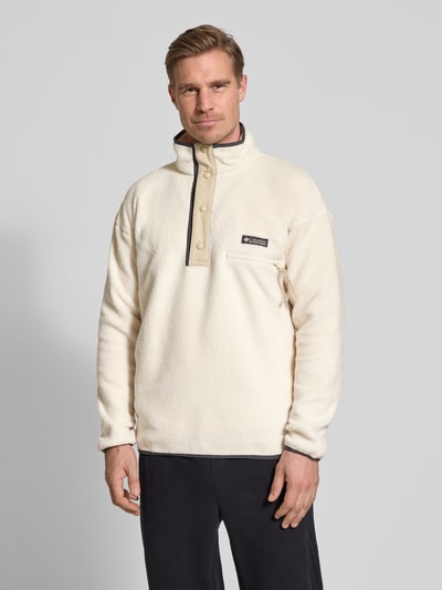 Męski model nosi Fleecepullover mit Stehkragen und Knopfleiste von Columbia, krój regular fit, wyhaftowane logo, kieszeń zapinana na zamek błyskawiczny, długość tyłu w rozmiarze M 73 cm, długość rękawów w rozmiarze M 70 cm, jasny kolor, gładka tekstura. Ganzkörperansicht z przodu.