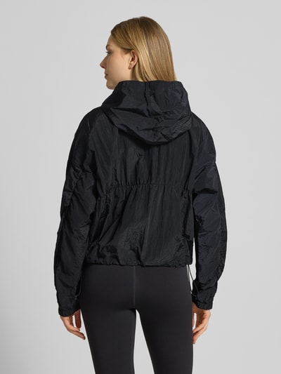 Frauenmodel trägt Jacke mit Kapuze und Logo von Under Armour in Schwarz, Regular-Fit, strukturierte Oberfläche, Ganzkörperansicht Rückansicht, Rückenlänge bei Größe S 57 cm, Schulterbreite 66 cm, Brustweite 64 cm, Ärmellänge 51 cm.