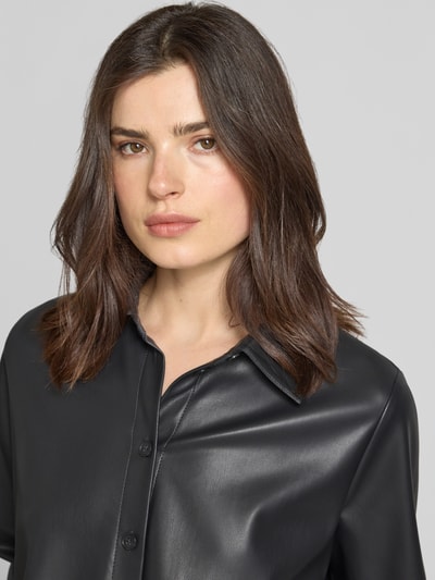 Modelka nosi Relaxed Fit Hemdjacke in Leder-Optik Modell 'Zeather' od Someday w czerni, z wykładanym kołnierzem i listwą guzikową, damska kurtka, imitacja skóry, długość tyłu w rozmiarze 36: 52 cm, szerokość ramion w rozmiarze 36: 40 cm, szerokość klatki piersiowej w rozmiarze 36: 65 cm, długość rękawów w rozmiarze 36: 63 cm, gładka faktura; zbliżenie na górną część ciała z przodu.