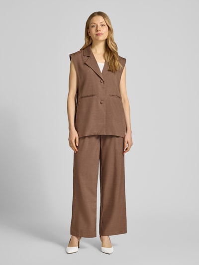 Frauenmodel trägt braune Stoffhose mit gelegten Falten von Little Mistress, Wide Leg, lockerer Schnitt, strukturierte Oberfläche, Ganzkörperansicht von vorne.