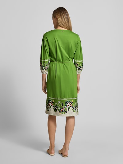 Frauenmodel trägt Knielanges Kleid mit Bindegürtel von Marc Cain in Grün mit buntem Blumenmuster, reine Baumwolle, Rückansicht, Ganzkörperansicht.