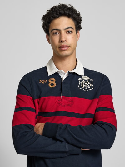 Männermodel trägt Regular Fit Rugby Shirt mit Logo- und Motiv-Stitching von Superdry in Dunkelblau mit roten Streifen, Reine Baumwolle, Polokragen, kurze Knopfleiste, Label-Patch, Rückenlänge bei Größe M 70 cm, Nahaufnahme Oberkörper.