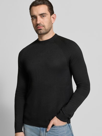 Męski model nosi Regular Fit Strickpullover mit Woll-Anteil von BOSS Green, czarny, dzianinowa struktura, nadruk z logo, okrągły dekolt, długie rękawy, ściągacze przy mankietach i na dole, optyka jednokolorowa, krój regular fit, widok całej sylwetki z przodu.