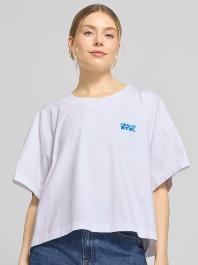 Frauenmodel trägt Damen T-Shirt mit überschnittenen Schultern Modell 'GIXY' von American Vintage aus reiner Baumwolle, Logo-Print, lockere Passform, Rundhalsausschnitt, weiße Farbe, Ganzkörperansicht von vorne.