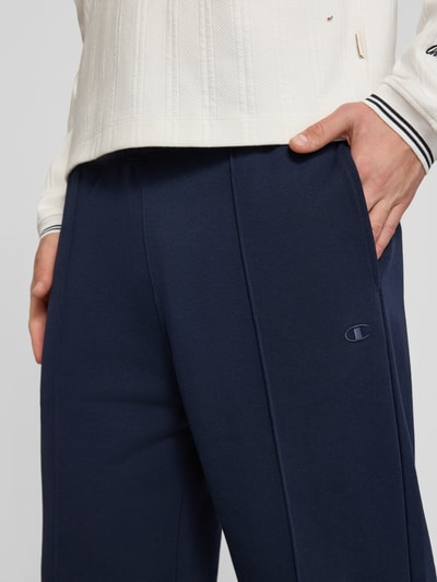 Männermodel trägt Sweatpants mit Längsnähten und Label-Stitching von CHAMPION, mieszanka bawełny, krój relaxed fit, elastyczny pas, ciemnoniebieski kolor, wyhaftowane logo, długość wewnętrzna nogawki w rozmiarze M: 81 cm, zbliżenie na przednią część spodni, ujęcie detailaufnahme.