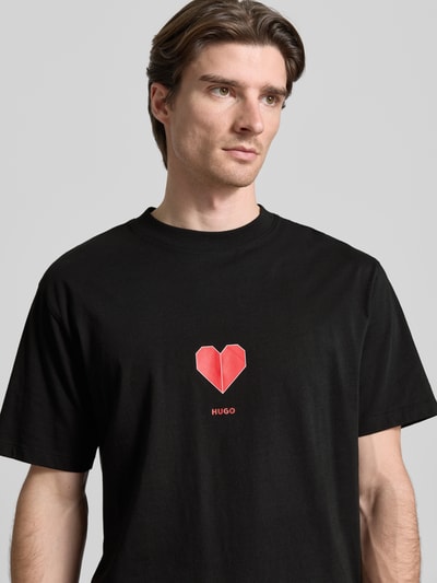 Männermodel trägt Regular Fit T-Shirt aus reiner Baumwolle Modell 'DALENTIN' von HUGO in Schwarz mit rotem Herzprint, Nahaufnahme Oberkörper.