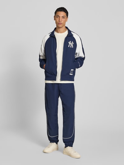 Männermodel trägt Relaxed Fit Tracksuit mit Logo-Patch von Champion, blau mit weißen Details, elastische Abschlüsse, Ganzkörperansicht von vorne.