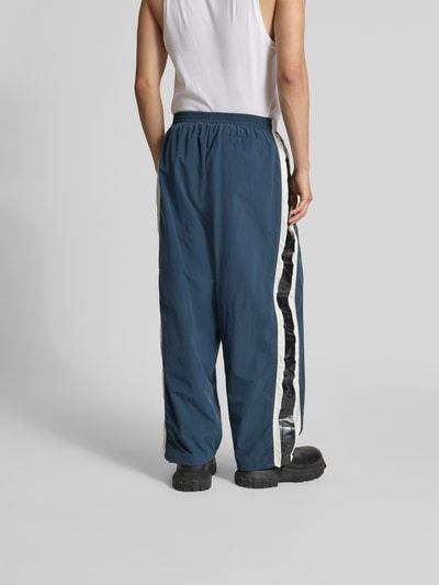 Männermodel trägt Trackpants mit elastischem Bund von VETEMENTS in Blau mit Galonstreifen, Regular Fit, Mesh-Futter, Ganzkörperansicht Rückansicht.