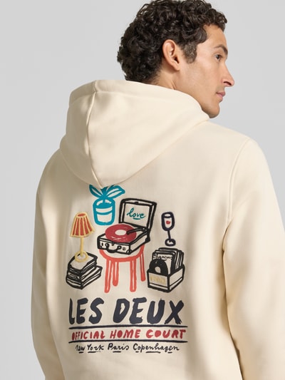 Männermodel trägt Hoodie aus reiner Baumwolle mit Logo-Print Modell 'Record' von Les Deux, Cremefarbe, Rückansicht, Ganzkörperansicht. Regular Fit, Kapuze mit Tunnelzug, langärmlig, gerippte Abschlüsse, Kängurutasche, farbenfroher Print mit Schriftzügen und grafischen Elementen, Rückenlänge 70 cm, Ärmellänge 74 cm.