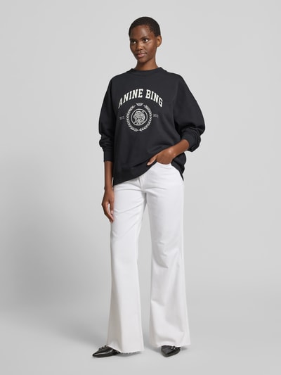 Frauenmodel trägt Oversized Fit Sweatshirt mit Logo-Print von Anine Bing aus Reine Baumwolle, schwarzer Stoff, weißer Print, elastische Abschlüsse, Rundhalsausschnitt, Rückenlänge 61 cm bei Größe S, Ganzkörperansicht von vorne.