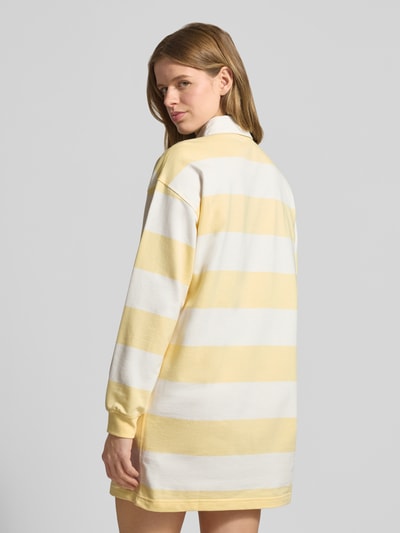 Frauenmodel trägt Regular Fit Minikleid mit Polokragen Modell 'Varsity' von Calvin Klein Jeans aus reiner Baumwolle, langärmelig, gestreiftes gelb-weißes Design, Logo-Stitching, elastische Ärmelabschlüsse, Rückenlänge bei Größe S 84 cm, Brustweite 52 cm, Ärmellänge 53 cm, Ganzkörperansicht Rückansicht.