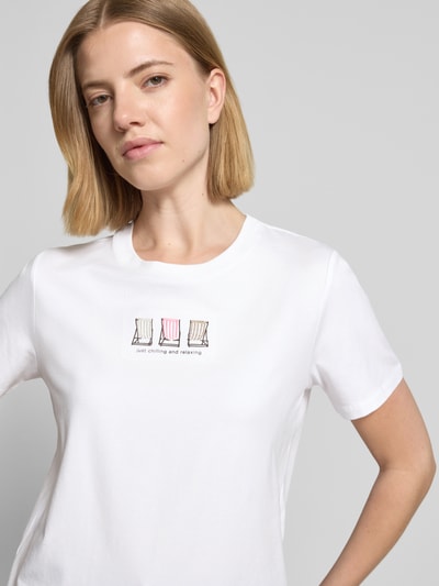 Modelka kobieca ma na sobie T-Shirt z okrągłym dekoltem od comma, bawełniany miks z dodatkiem modalu, krój regular fit, białe, ze statement printem i ozdobnymi przeszyciami, długość tyłu w rozmiarze 36 to 49 cm, zbliżenie na górną część ciała z przodu.