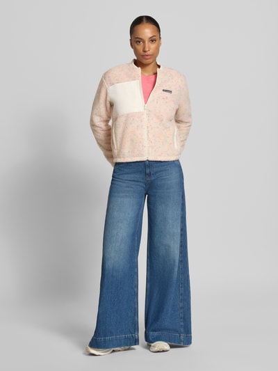 Modelka kobieca ma na sobie Regular Fit Jacke mit Stehkragen od Columbia, kolor kremowy z kolorowymi akcentami, tekstura polaru, logo-patch, kieszenie na piersi i boczne, zamek błyskawiczny, krój regular fit, długość pleców w rozmiarze S: 49 cm, szerokość ramion w rozmiarze S: 44 cm, szerokość klatki piersiowej w rozmiarze S: 50 cm, długość rękawa w rozmiarze S: 61 cm, widok całej sylwetki z przodu.